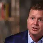 Man in a blue blazer speaks during a discussion, with a blurred background.
