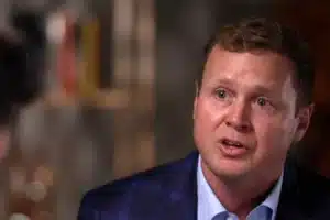 Man in a blue blazer speaks during a discussion, with a blurred background.