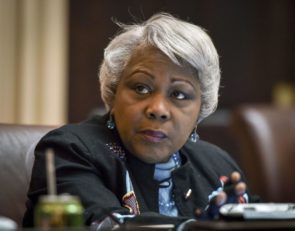 Close-up of a Black woman with short gray hair, looking to her left, wearing turquoise earrings and a dark blazer.