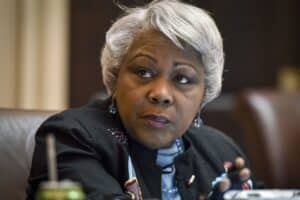 Close-up of a Black woman with short gray hair, looking to her left, wearing turquoise earrings and a dark blazer.