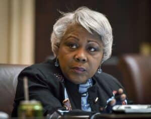 Close-up of a Black woman with short gray hair, looking to her left, wearing turquoise earrings and a dark blazer.