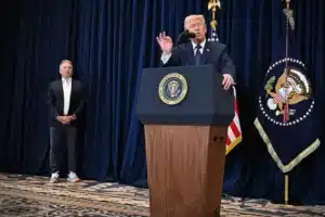 A man speaks at a podium bearing the presidential seal, with an aide standing to the left on a dark-curtain stage behind flags.