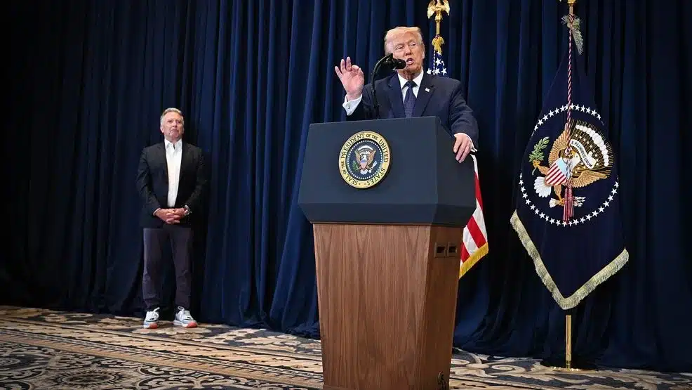 A man speaks at a podium bearing the presidential seal, with an aide standing to the left on a dark-curtain stage behind flags.