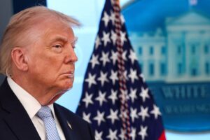 Portrait of former President Donald Trump in a dark suit and light blue tie, with an American flag and government building backdrop.
