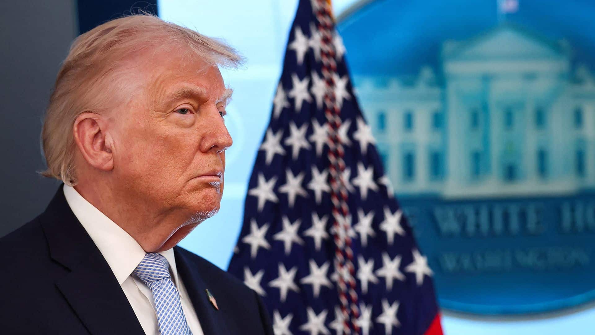 Portrait of former President Donald Trump in a dark suit and light blue tie, with an American flag and government building backdrop.