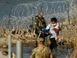 Military officer assisting a woman and a child as they wade through water near a barbed-wire barrier, offering protection and support.