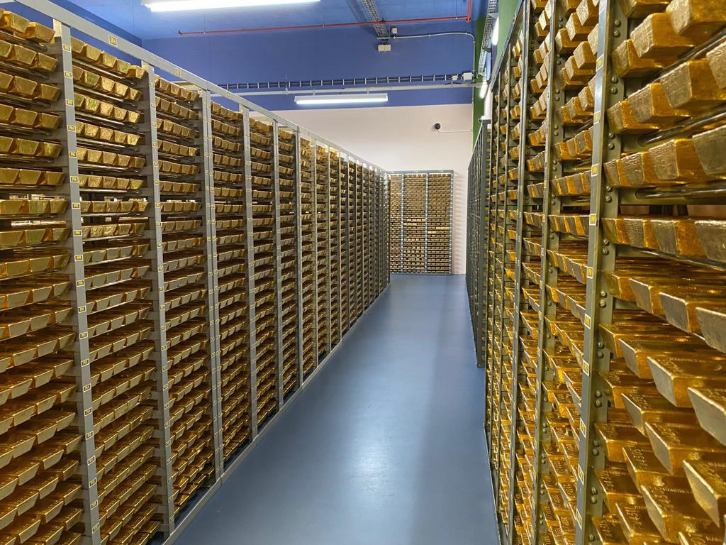 Corridor of metal racks filled with neatly stacked gold bars in a secure storage vault, viewed from one end.
