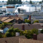 Residential neighborhood with brown shingle roofs in the foreground and an industrial complex with smokestacks in the background.