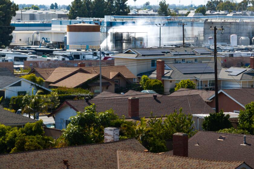 Residential neighborhood with brown shingle roofs in the foreground and an industrial complex with smokestacks in the background.
