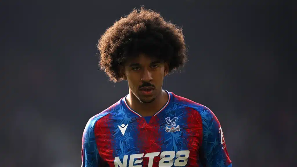 Young male footballer with big curly hair wearing a blue and red Crystal Palace jersey, looking intently ahead on the pitch.