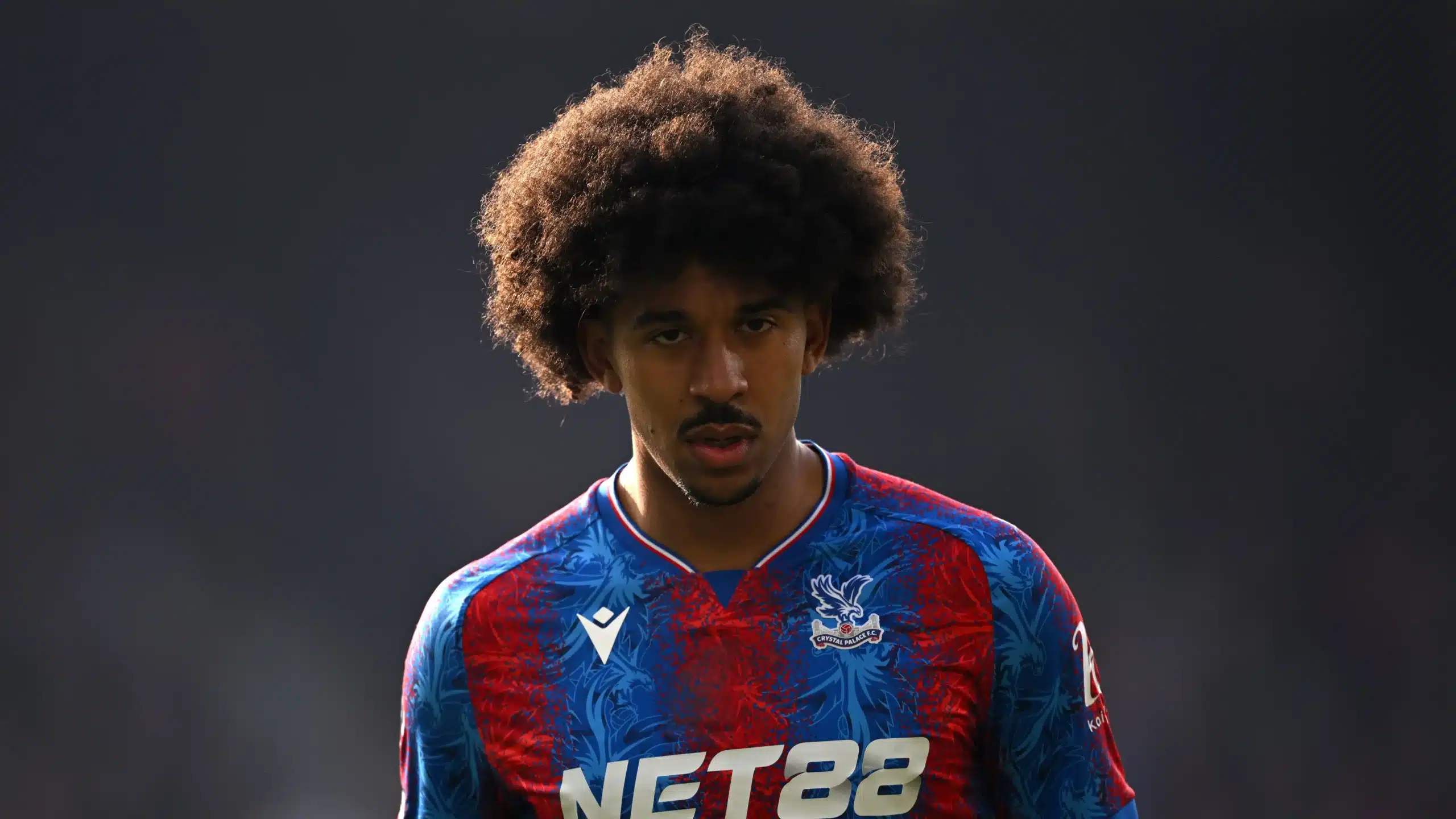 Young male footballer with big curly hair wearing a blue and red Crystal Palace jersey, looking intently ahead on the pitch.