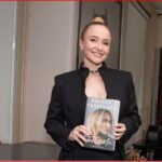 Woman in a black blazer smiles while holding a hardcover book with a blonde model on the cover.