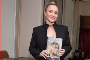 Woman in a black blazer smiles while holding a hardcover book with a blonde model on the cover.