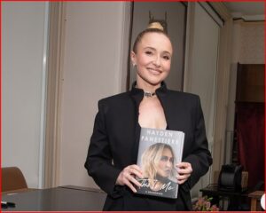 Woman in a black blazer smiles while holding a hardcover book with a blonde model on the cover.