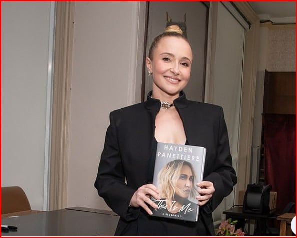 Woman in a black blazer smiles while holding a hardcover book with a blonde model on the cover.