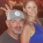 Smiling man wearing a gray baseball cap and a woman in a blue sleeveless dress posing together at a social event.