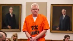 Man in an orange prison jumpsuit stands at the defendant's stand in a courtroom, with framed portraits on the wall behind him.