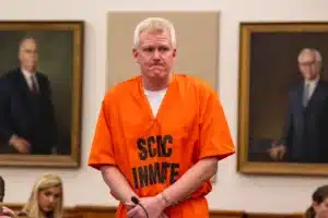Man in an orange prison jumpsuit stands at the defendant's stand in a courtroom, with framed portraits on the wall behind him.