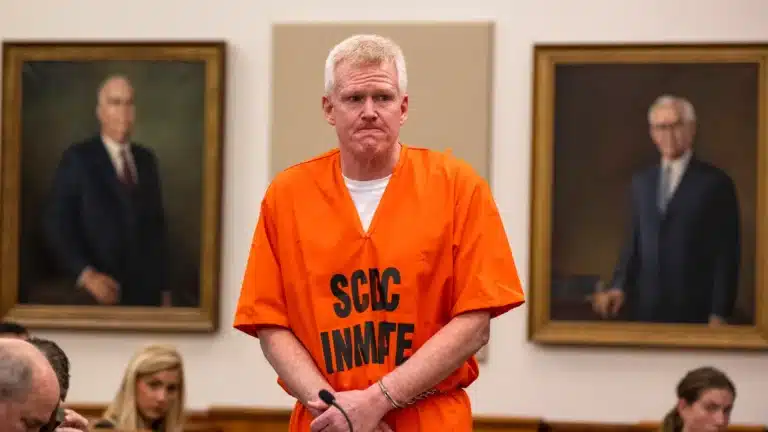 Man in an orange prison jumpsuit stands at the defendant's stand in a courtroom, with framed portraits on the wall behind him.