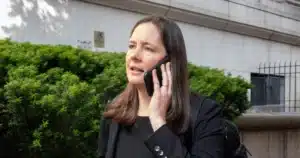 Woman in a black blazer talking on a cellphone outdoors, next to a green hedge and a stone building background.