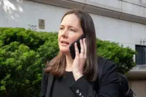Woman in a black blazer talking on a cellphone outdoors, next to a green hedge and a stone building background.