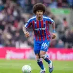 Soccer player in blue and red striped kit dribbling a ball on a green pitch with a crowd in the background.