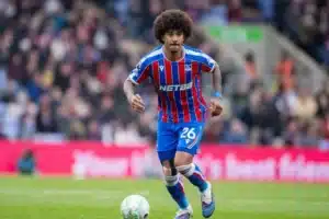 Soccer player in blue and red striped kit dribbling a ball on a green pitch with a crowd in the background.