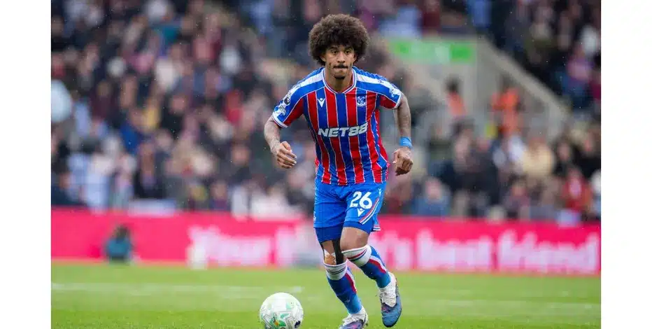 Soccer player in blue and red striped kit dribbling a ball on a green pitch with a crowd in the background.