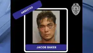 Mugshot of a man with short dark hair, a blue 'ARRESTED' banner, and a name plate reading 'JACOB BAKER' at the bottom.