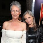 Two smiling women posing on a red carpet; one in a white off‑the‑shoulder dress with short gray hair, the other in a black leather jacket with long gray hair.