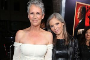 Two smiling women posing on a red carpet; one in a white off‑the‑shoulder dress with short gray hair, the other in a black leather jacket with long gray hair.