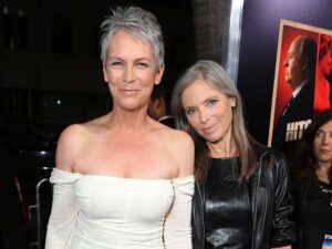 Two smiling women posing on a red carpet; one in a white off‑the‑shoulder dress with short gray hair, the other in a black leather jacket with long gray hair.
