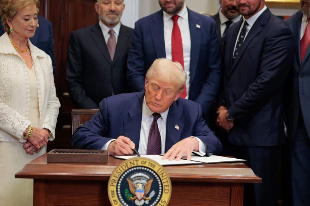 Donald Trump signs a document at a desk while a woman in white and several suited men look on, the presidential seal in the foreground.