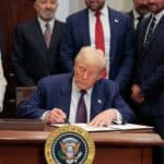 Donald Trump signs a document at a desk while a woman in white and several suited men look on, the presidential seal in the foreground.