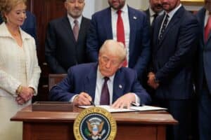 Donald Trump signs a document at a desk while a woman in white and several suited men look on, the presidential seal in the foreground.