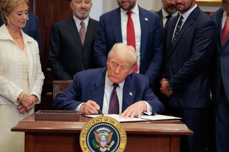 Donald Trump signs a document at a desk while a woman in white and several suited men look on, the presidential seal in the foreground.