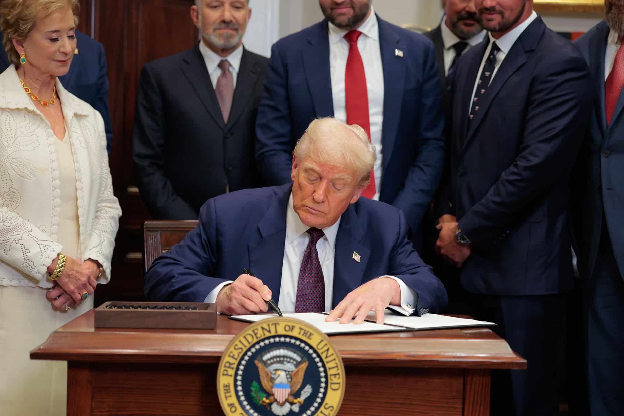 Donald Trump signs a document at a desk while a woman in white and several suited men look on, the presidential seal in the foreground.