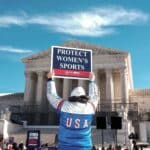 Person in a USA jersey raises a sign that reads 'Protect Women’s Sports' at a rally outside a neoclassical building, with a crowd in the background.