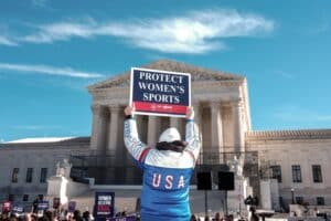 Person in a USA jersey raises a sign that reads 'Protect Women’s Sports' at a rally outside a neoclassical building, with a crowd in the background.