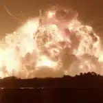 Nighttime rocket launch pad explosion with a massive orange fireball and thick smoke rising over the facility.