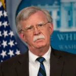 Senior man with gray hair and mustache, in a dark suit and striped tie, speaking at a White House press briefing with an American flag nearby.
