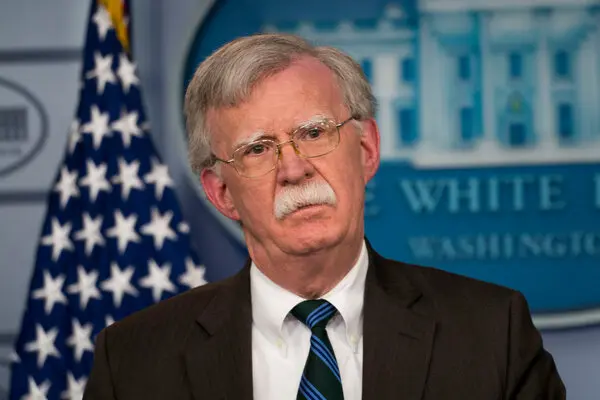 Senior man with gray hair and mustache, in a dark suit and striped tie, speaking at a White House press briefing with an American flag nearby.