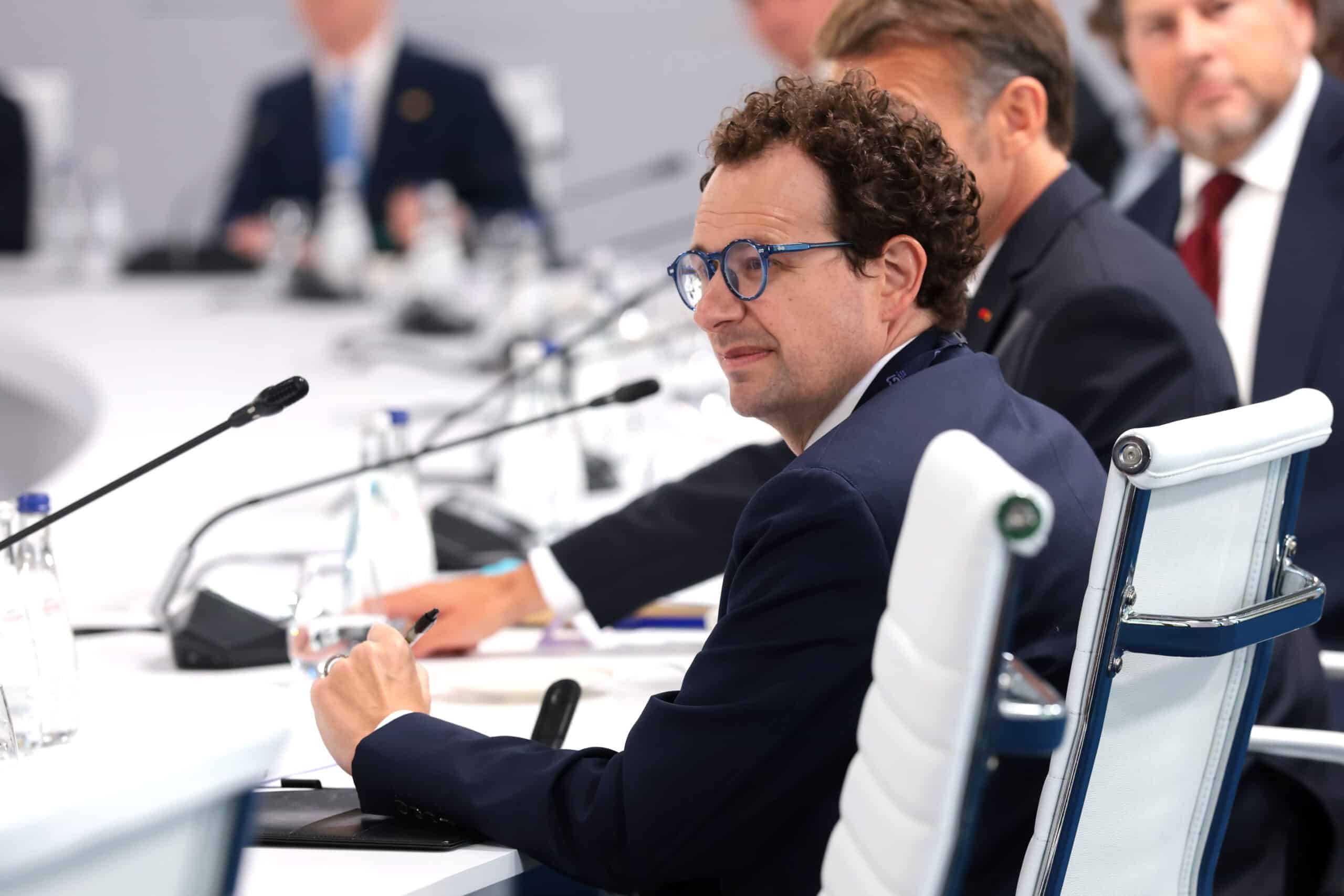 Man in a suit and blue glasses signs a document at a conference table, others blurred in the background.