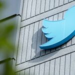 Blue Twitter bird logo mounted on a gray brick exterior wall, viewed from below with foreground leaves partially framing the image.