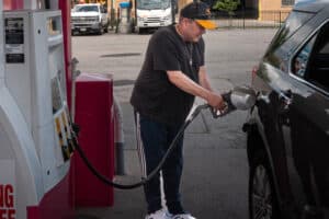 Man in a black t-shirt and orange cap refueling a dark gray SUV at a gas pump.