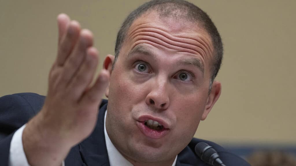 Man in suit speaking into a microphone while gesturing with an open hand, as if making a point during a discussion