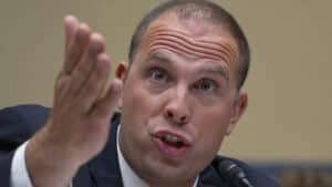Man in suit speaking into a microphone while gesturing with an open hand, as if making a point during a discussion
