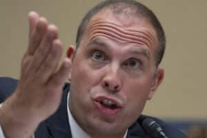 Man in suit speaking into a microphone while gesturing with an open hand, as if making a point during a discussion