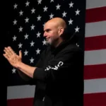 Speaker with a shaved head and gray beard in a black hoodie claps on stage in front of a large American flag backdrop.