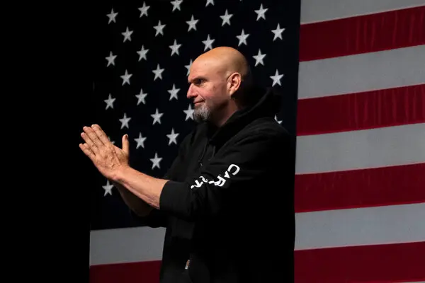 Speaker with a shaved head and gray beard in a black hoodie claps on stage in front of a large American flag backdrop.